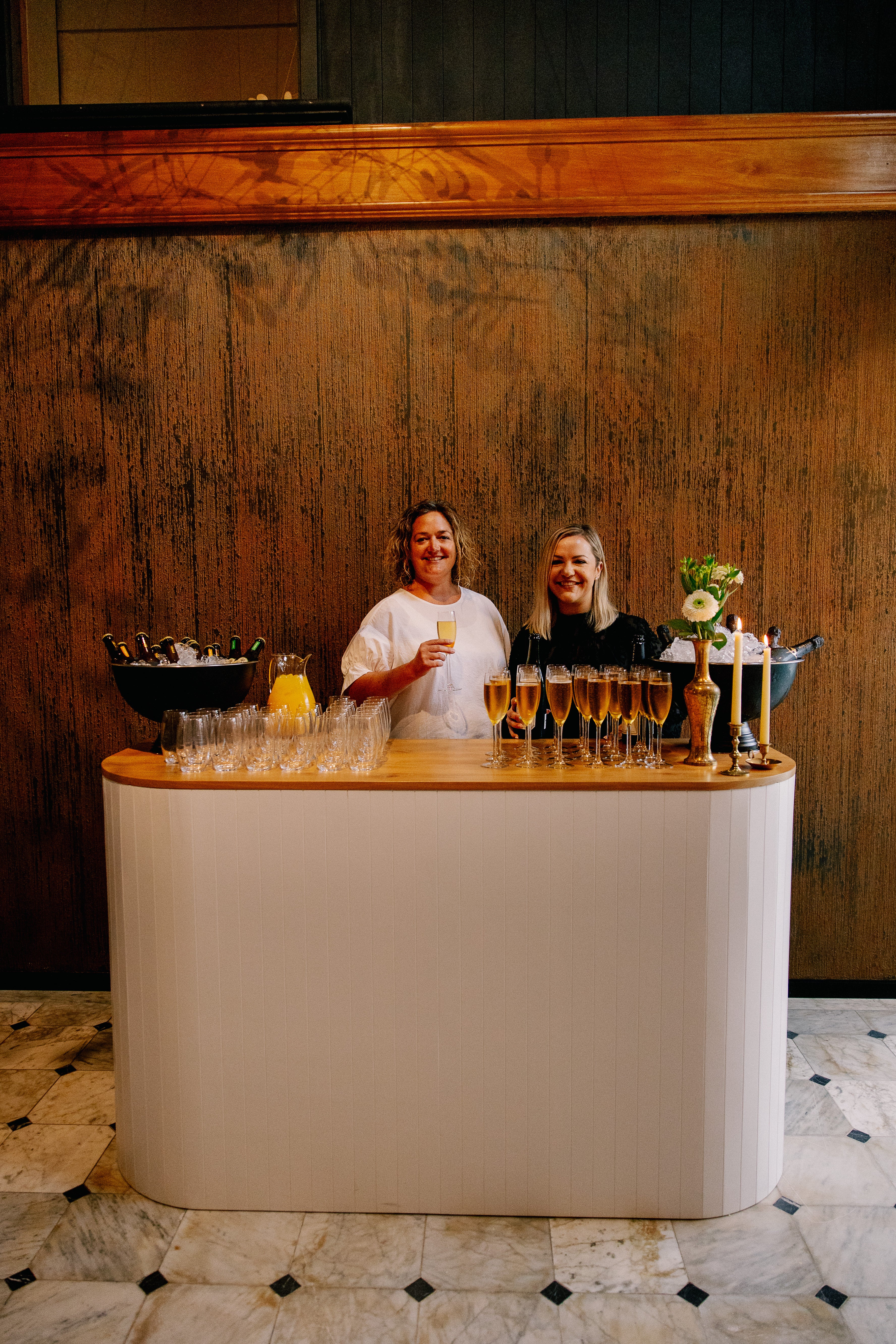 Two bartenders behind a bar counter with drinks and glasses in a dimly lit room.